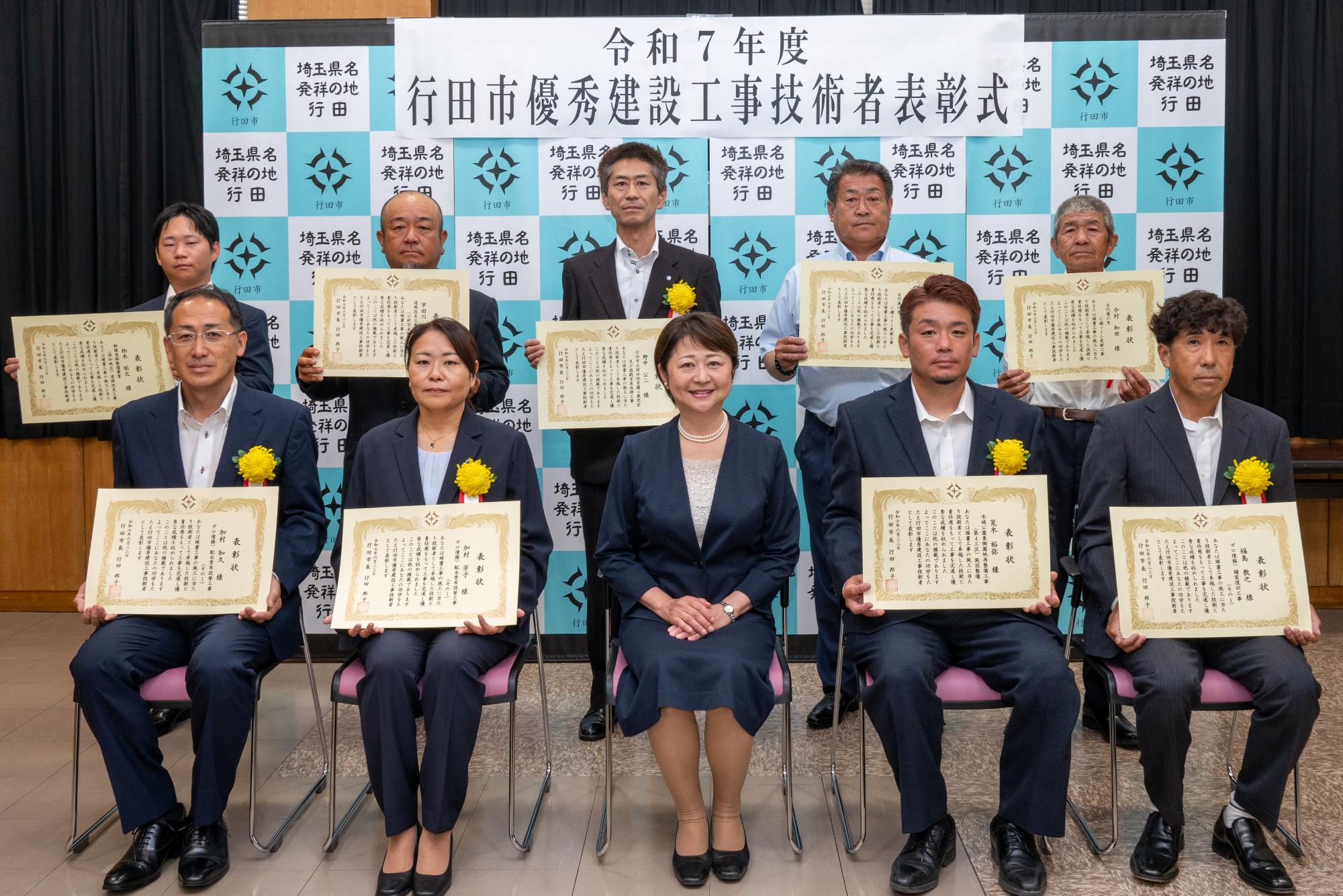 表彰式にて胸に黄色い花を付けたスーツ姿の表彰者8名が賞状を持って行田市長と一緒に並んでいる写真