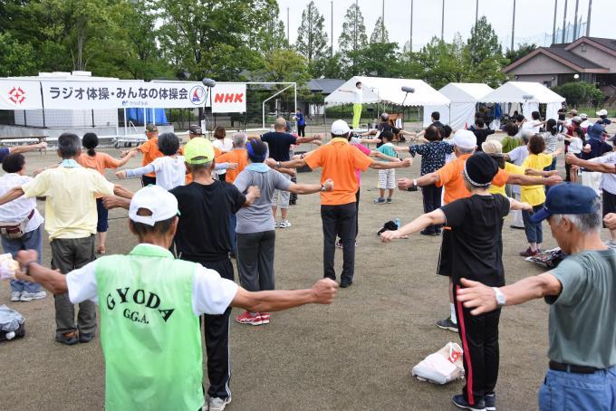 朝礼台に上がって体操するラジオ体操指導員と同様に、たくさんの参加者がラジオ体操を行なっている様子の写真