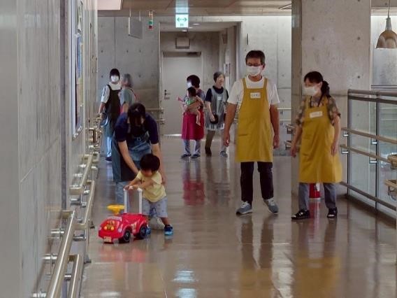 廊下で、おもちゃの車を押している1人の子どもを、世話をする中学生。その様子を二人の男女の民生委員が見守っている様子。