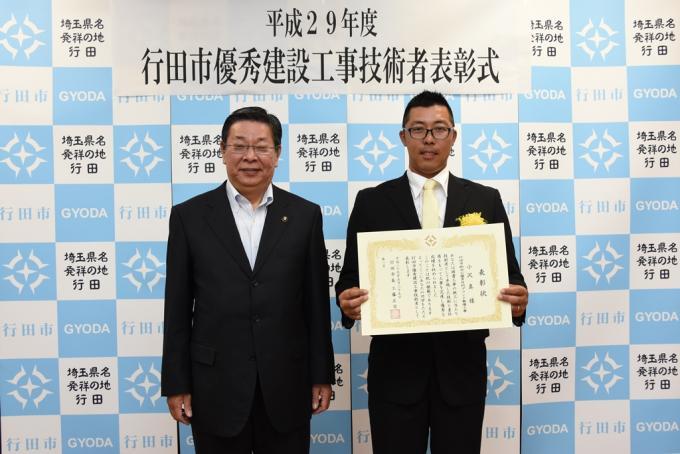 表彰式にて胸に黄色い花を付けたスーツ姿の小沢さんが賞状を持って行田市長と一緒に記念撮影している写真