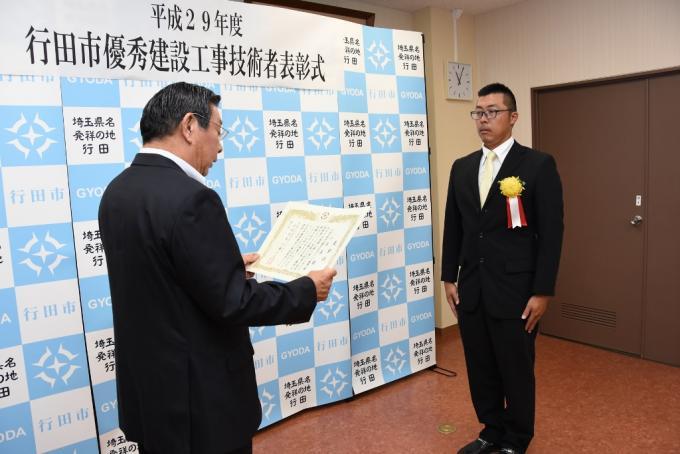 表彰式にて賞状を読み上げている行田市長の前に胸に黄色い花を付けたスーツ姿の小沢さんが立っている写真