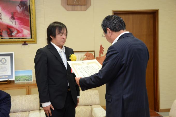表彰式にて胸に黄色い花を付けたスーツ姿の小林さんが行田市長より表彰されている写真