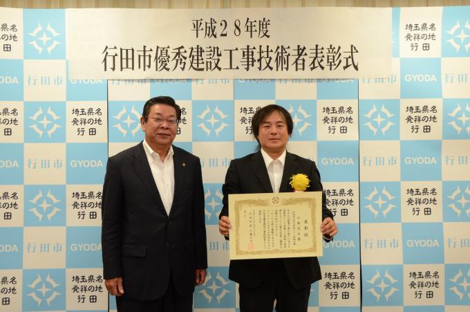 表彰式にて胸に黄色い花を付けたスーツ姿の小林さんが賞状を持って行田市長と一緒に記念撮影している写真