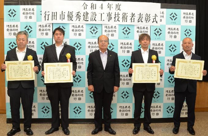 表彰式にて胸に黄色い花を付けたスーツ姿の表彰者4名が賞状を持って石井市長と一緒に並んで立っている写真