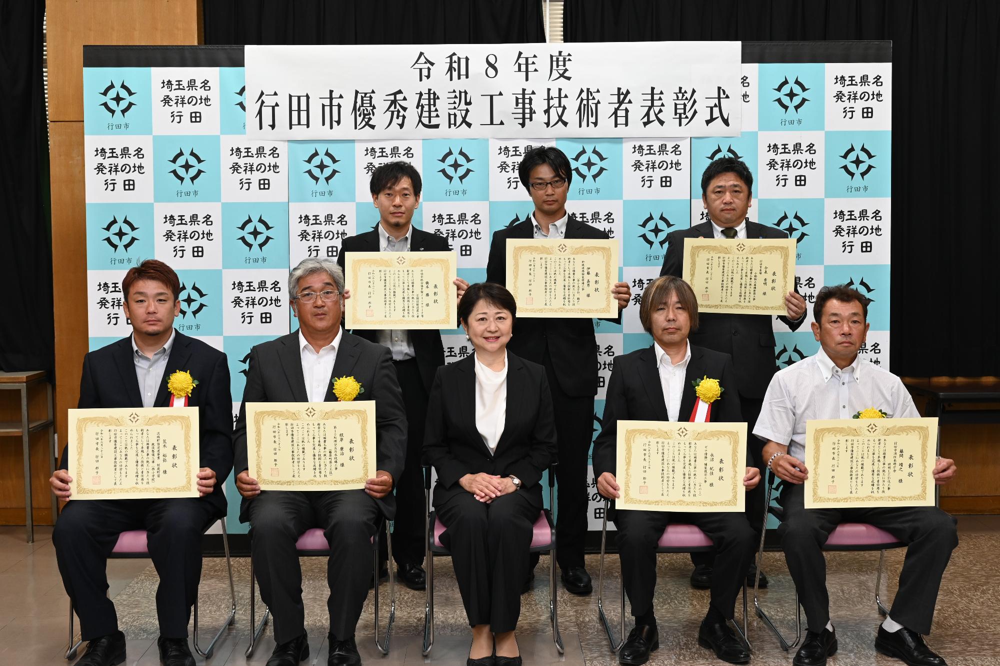 表彰式にて胸に黄色い花を付けたスーツ姿の表彰者7名が賞状を持って行田市長と一緒に並んでいる写真