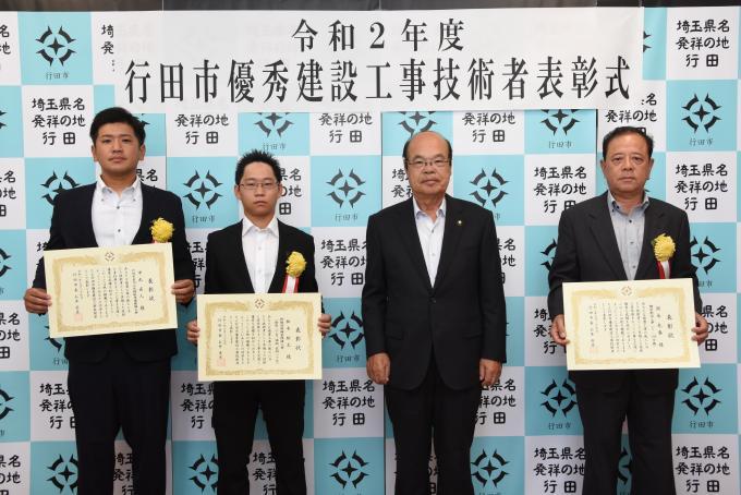 表彰式にて胸に黄色い花を付けたスーツ姿の表彰者3名が賞状を持ち、石井市長と一緒に記念撮影している写真