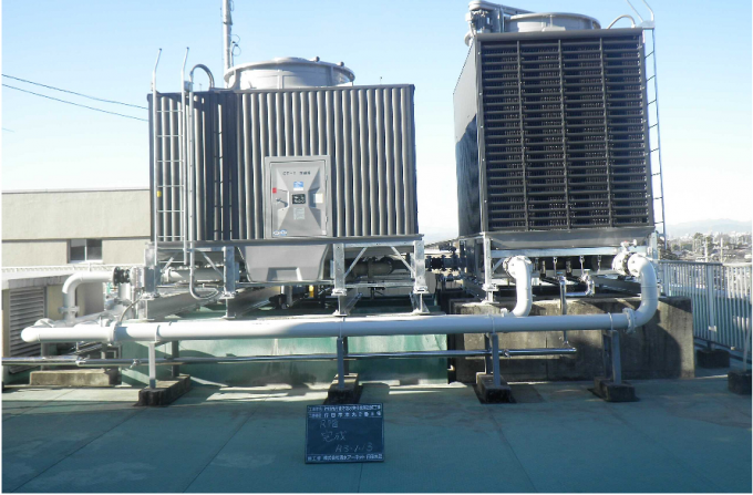 工事現場用黒板と屋上に設置されている、冷温水発生機の配管と二つのタンクの写真