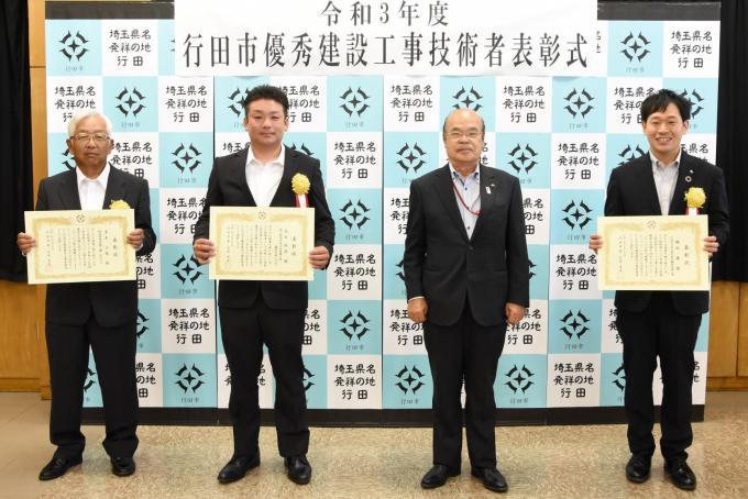表彰式にて胸に黄色い花を付けたスーツ姿の表彰者3名が賞状を持って石井市長と一緒に並んで立っている写真