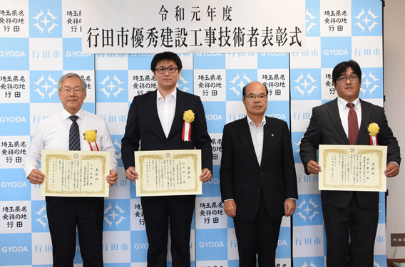 表彰式にて胸に黄色い花を付けた表彰された三田さん、杉山さん、根岸さんが賞状を持って石井市長と一緒に記念撮影している写真