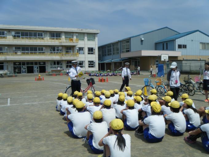 生徒たちの前に立って話をしている交通指導員と運動場に座って話を聞いている体操服に黄色の帽子を被った生徒たちの写真