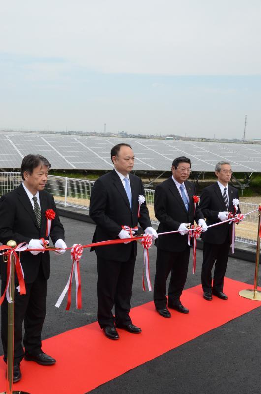 行田ソーラーウェイ太陽光発電所の竣工式でスーツ姿で胸章を付けた男性4人が赤い毛氈の上に立ってテープカットを行っている様子の写真