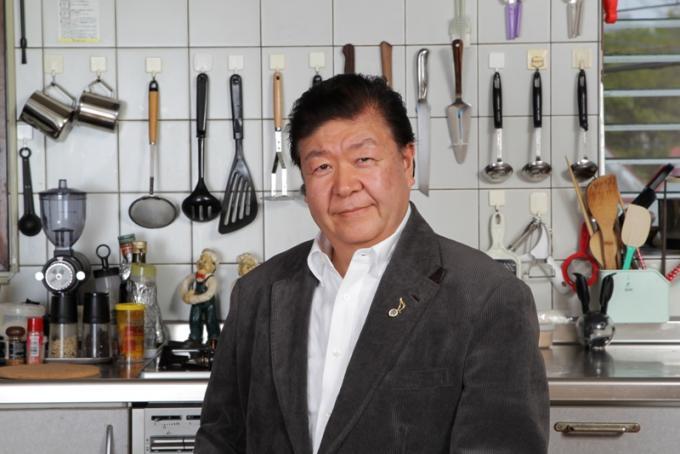 お玉やフライ返しなどの調理器具がたくさん置かれている調理場を背景に写っている田中利幸さんの写真
