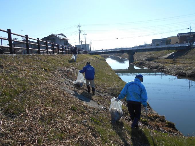 ごみ袋と火ばさみを持って川沿いの土手を歩きながらごみを拾っている清掃活動の参加者の写真