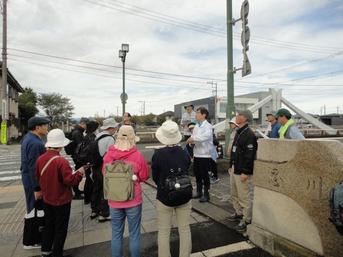 忍川遊歩道にてリュックやバックを持ち帽子を被った参加者の人々が白いジャンパーを着た男性の周りに集まり、解説を聞いている写真