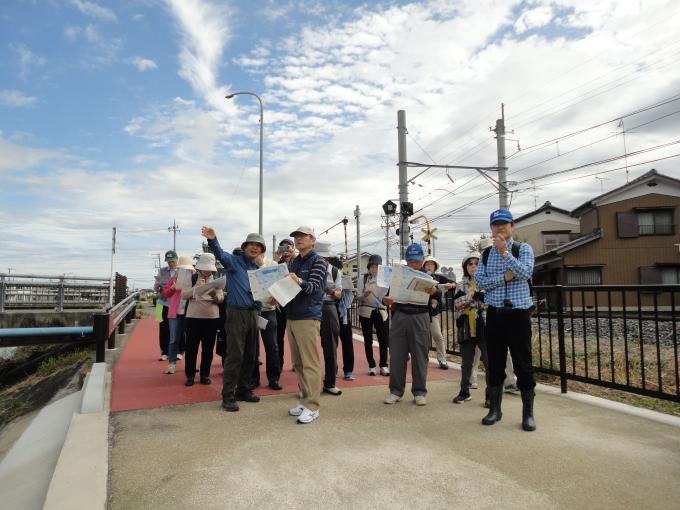 青いジャンパーを着た男性が前方を左手で指し示し、指示された方向を見ている参加者と手に持っている資料を見て調べている様子の参加者達の写真