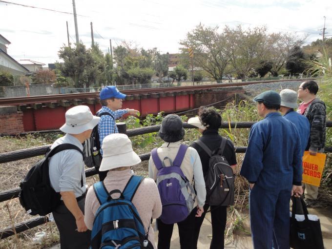 赤色の鉄道が見える場所に集まった参加者の方々が青い帽子を被った男性の説明を熱心に聞いている様子の写真