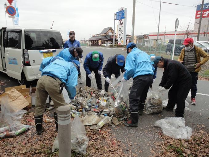参加者の方々が空き缶や落ち葉などの沢山のごみを道路端で広い集め、ゴミ袋に入れている写真