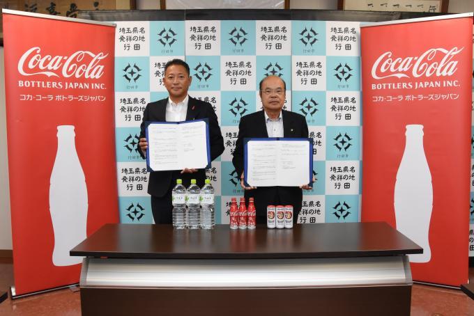 協定書を持っている市川様と石井市長がコカ・コーラ社製品が置かれた長机の後ろに立っている写真
