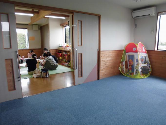 手前に青い絨毯か敷かれた部屋、引き戸で仕切られた奥にフローリングの部屋があり、親子が集まって円を描くように座っている写真