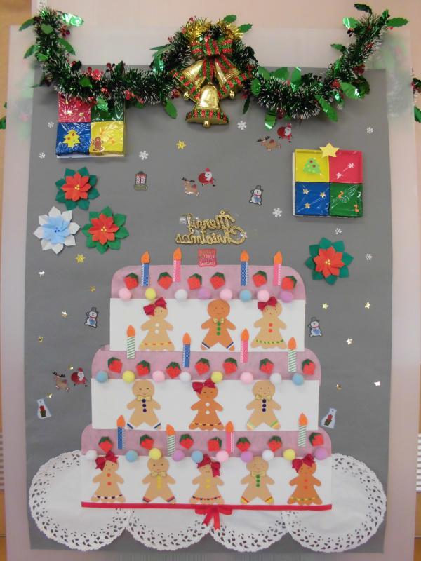 壁に飾られたクリスマスのリースとベル、人型のクッキーやろうそくが並んでいる折り紙や画用紙で作られたケーキのアート作品の写真