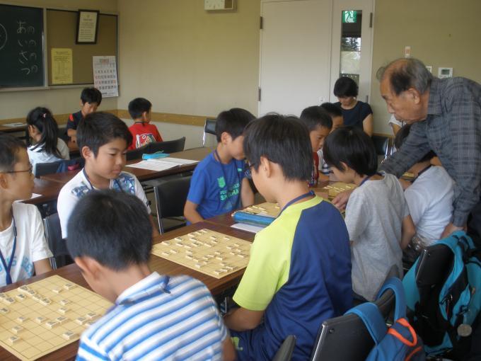 長机に1対1で向かい合って席につき、将棋を指している子供たちと将棋の駒を指さして指導をしている森先生の写真