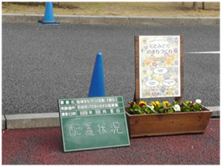 道路脇に「花とみどりのまちづくり」のポスターとその前には黄色や白色の花が咲いたプランターと配置状況を示した黒板が置いてある写真