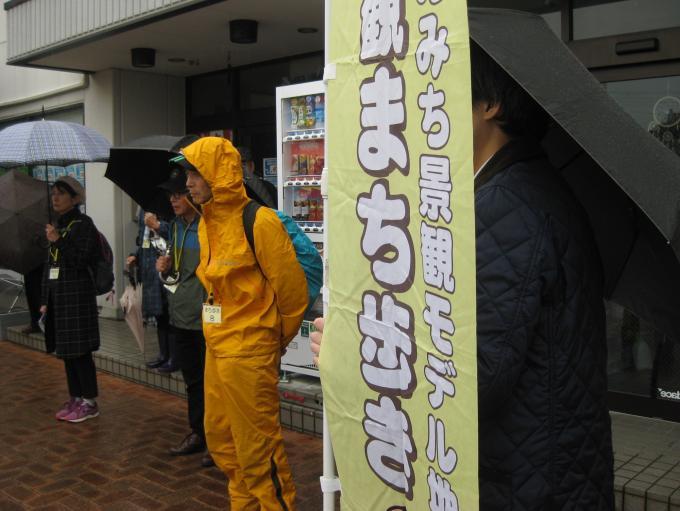 雨が降る中、「景観まち歩きののぼり」の周りに黄色のレインウェアを着た人や傘をさしている参加者の写真