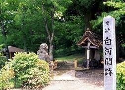 緑色の葉をつけた木々と奥につづく小道、右側に立っている「史跡白川関跡」と書かれた石碑の写真