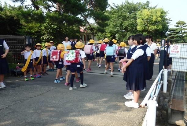埼玉小学校の入口の両脇に黄色いタスキをかけた小学生と埼玉中学校の生徒が立ち、登校中の小学生に挨拶をしている写真