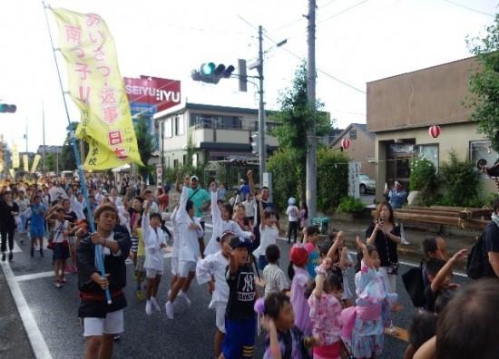 佐間天神社八坂祭にて、右手を挙げながら街の中心を歩いている南小学校と行田中学校の生徒たちと、その中の一人が黄色いのぼり旗を持って歩いている写真