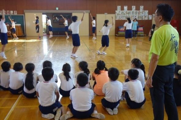 体育館で輪になり右手を挙げてだんべ踊りを踊っている行田中学校の生徒を南小学校の児童が座って見ている写真