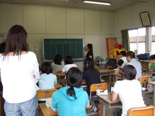 前方の黒板を使って、女性が授業をしており、子ども達が席に着いて授業を受けている様子を後ろから写している学習風景の写真