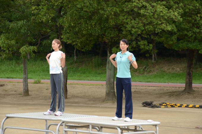 全国ラジオ体操連盟指導委員の女性二人が緑の木々の前の台の上に上がり、ラジオ体操の手本を見せている様子の写真