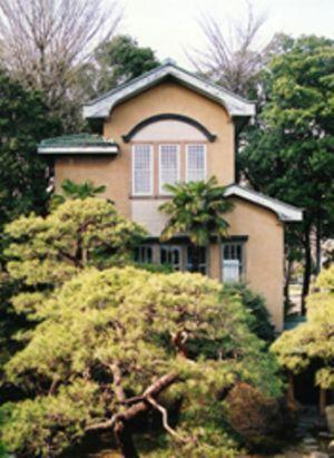 木々の奥に見えるベージュ色の三角屋根の建物で、縦長の窓が横にが3つ並んでいる洋館の外観写真