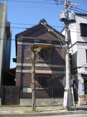 街路樹と電柱の奥に建っている、茶色いレンガ造りで2階建ての大澤家住宅旧文庫蔵の写真