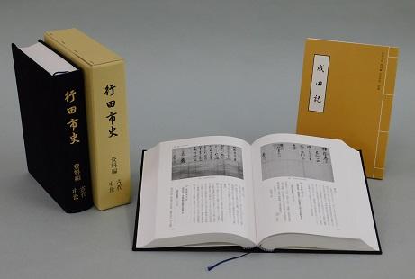 見開きになっている行田市史資料編古代中世の左側に「行田市史資料編 古代中世」の本と本のケースが立てて並べてあり、右側にオレンジ色の表紙の「成田記」の翻刻が立てて置いてある写真