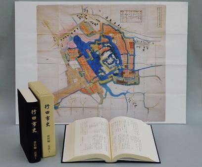 見開きになっている行田市史 資料編 近世1の左側に「行田市史 資料編 近世1」の本と本のケースが立てて並べて置いてあり、その後ろに忍城図が飾られている写真