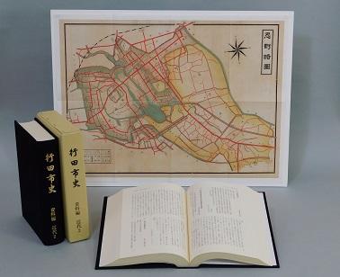 見開きになっている行田市史 資料編 近代2の左側に「行田市史 資料編 近代2」の本と本のケースが立てて並べて置いてある、その後ろに忍町略図が飾られている写真
