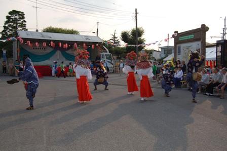 花が飾られた笠を頭に乗せた白い着物に赤い袴を着た3人の女性、その周りでお面を付けた獅子が舞を踊り、観衆の前で披露している写真