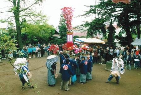 青い法被を着た男性らが花籠を高く持ち上げ、その周りで3頭の獅子が舞を踊っており、その様子を沢山の観衆が見ている写真